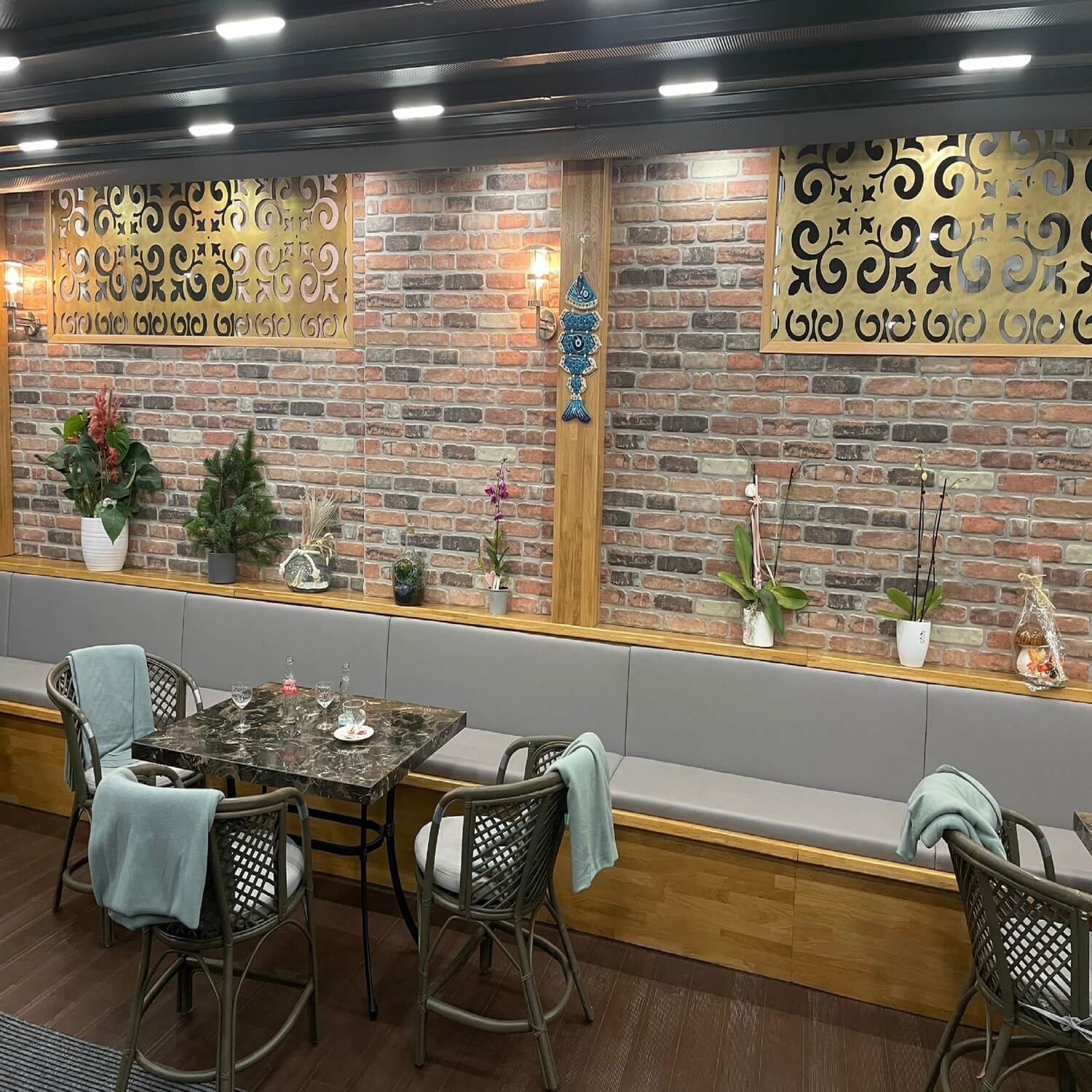 A restaurant or cafe seating area with Earthcore 3D faux brick wall panels, featuring built-in wooden benches with grey cushions, dining tables with wicker chairs and teal blankets, ornate gold decorative screens, various potted plants on a wooden shelf, and modern lighting